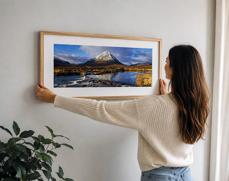 An oak-framed print of Buachaille Etive Mòr being hung on a plain wall